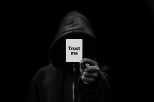 A man with a dark hood on and the word trust me on a piece of paper covering his face.